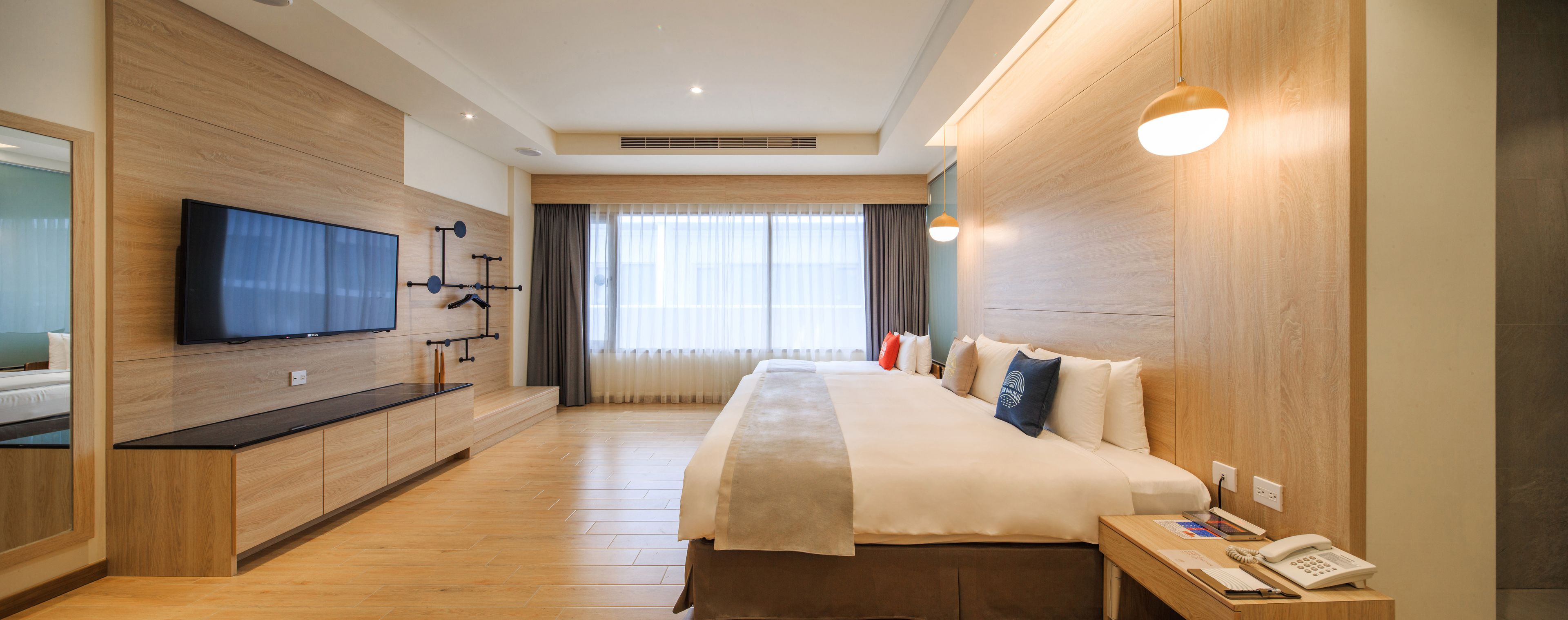 Quarto de hotel com uma cama grande, televisão de tela plana fixada na parede, um armário de madeira e uma mesinha de cabeceira com telefone.