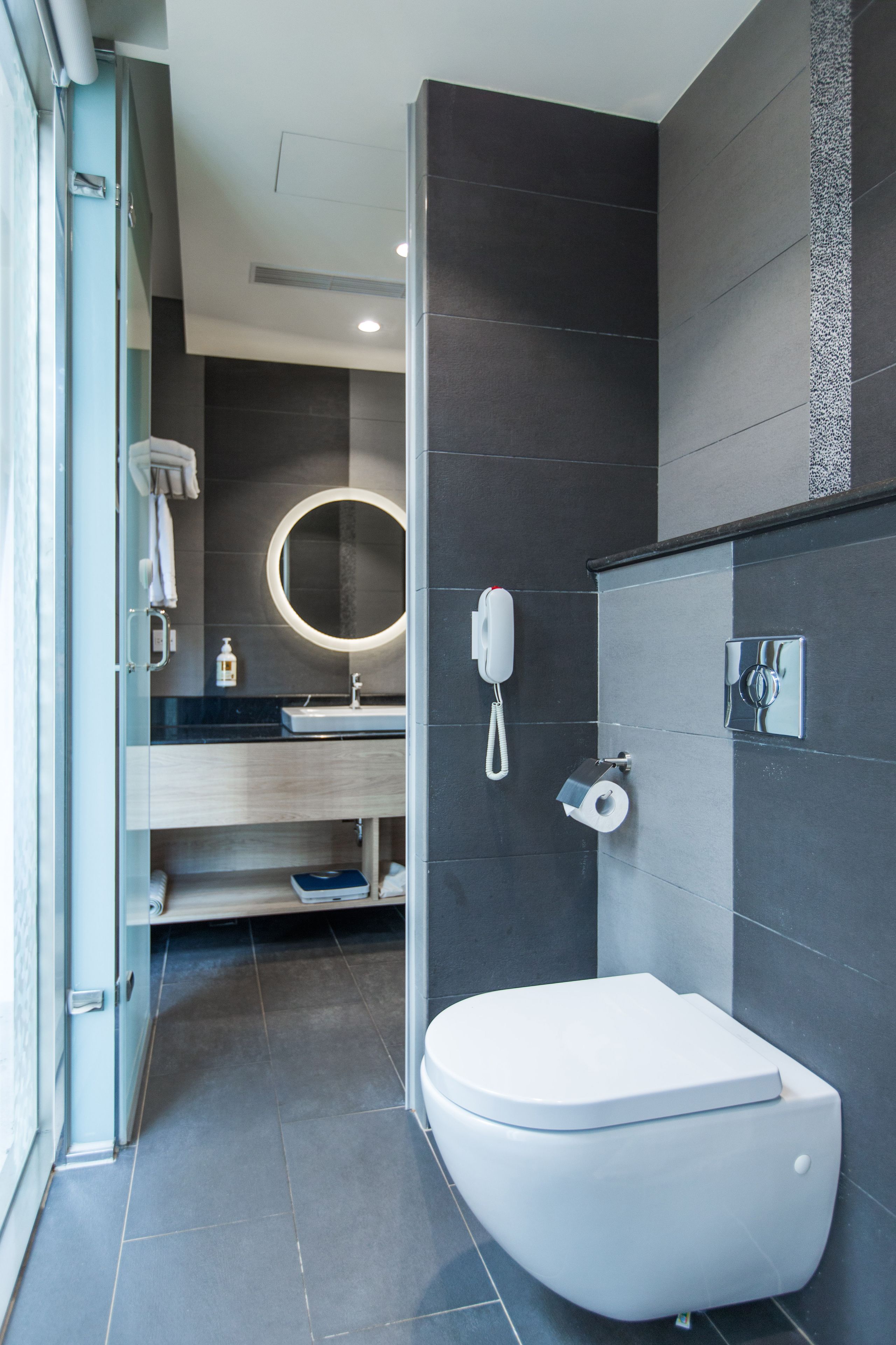 A modern bathroom with a white toilet, a round mirror, a handheld phone, and a towel rack.