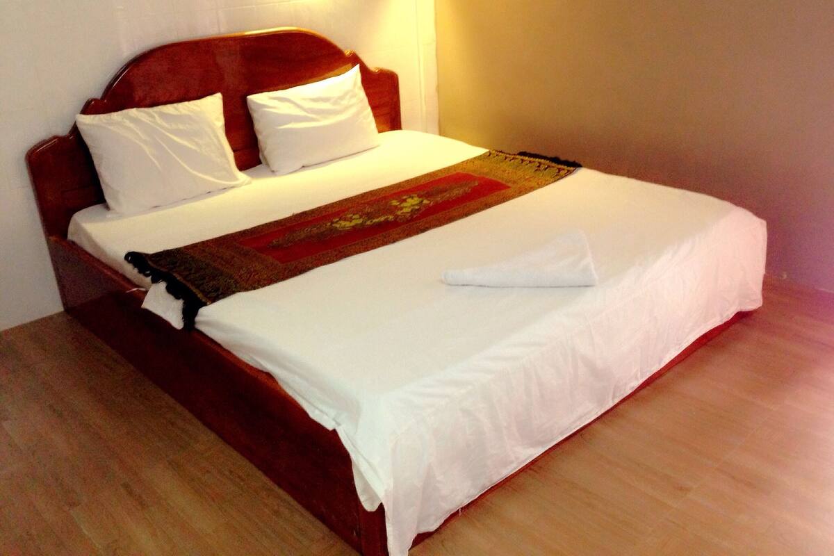 A neatly made bed with white linens and a brown headboard, a white pillow, and a folded white towel on the bed.