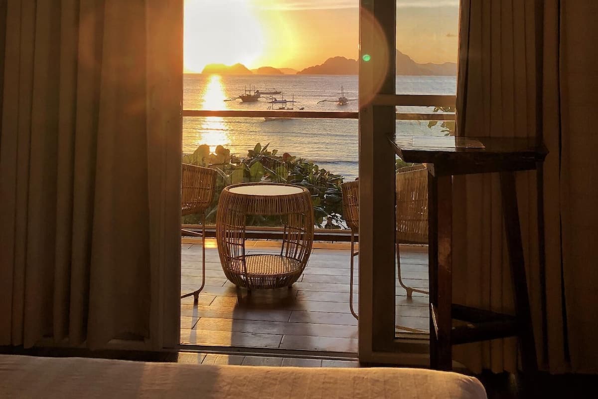 A room with a view of a sunset over the sea, a wicker chair, and a small table.