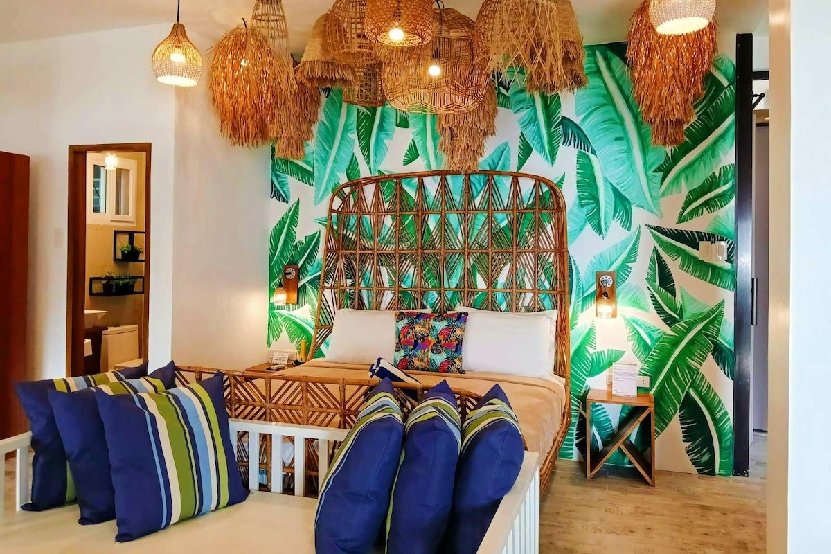 Een kamer met een bed, een houten hoofdbord, een muur met een tropisch bladpatroon en hangende rieten lampen.