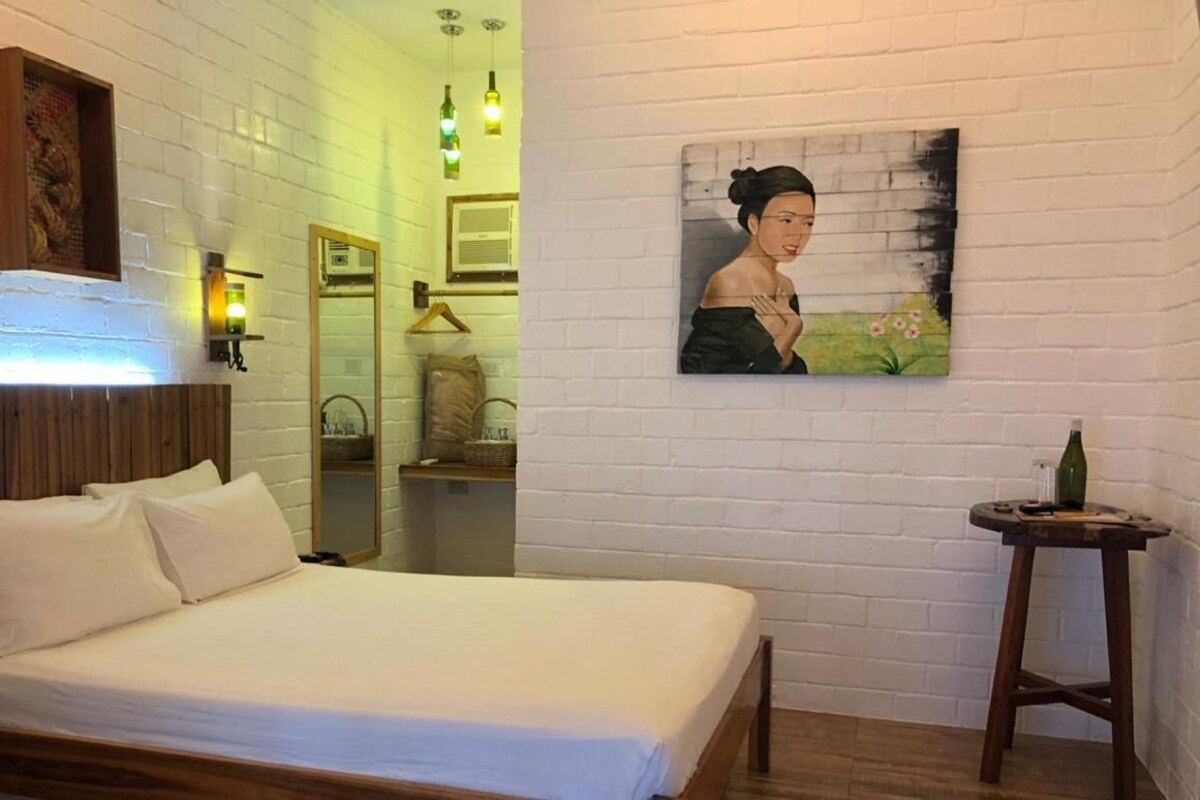 Een slaapkamer met een houten hoofdbord, een bed met witte lakens, een schilderij aan de muur, een klein krukje met een fles en een spiegel.