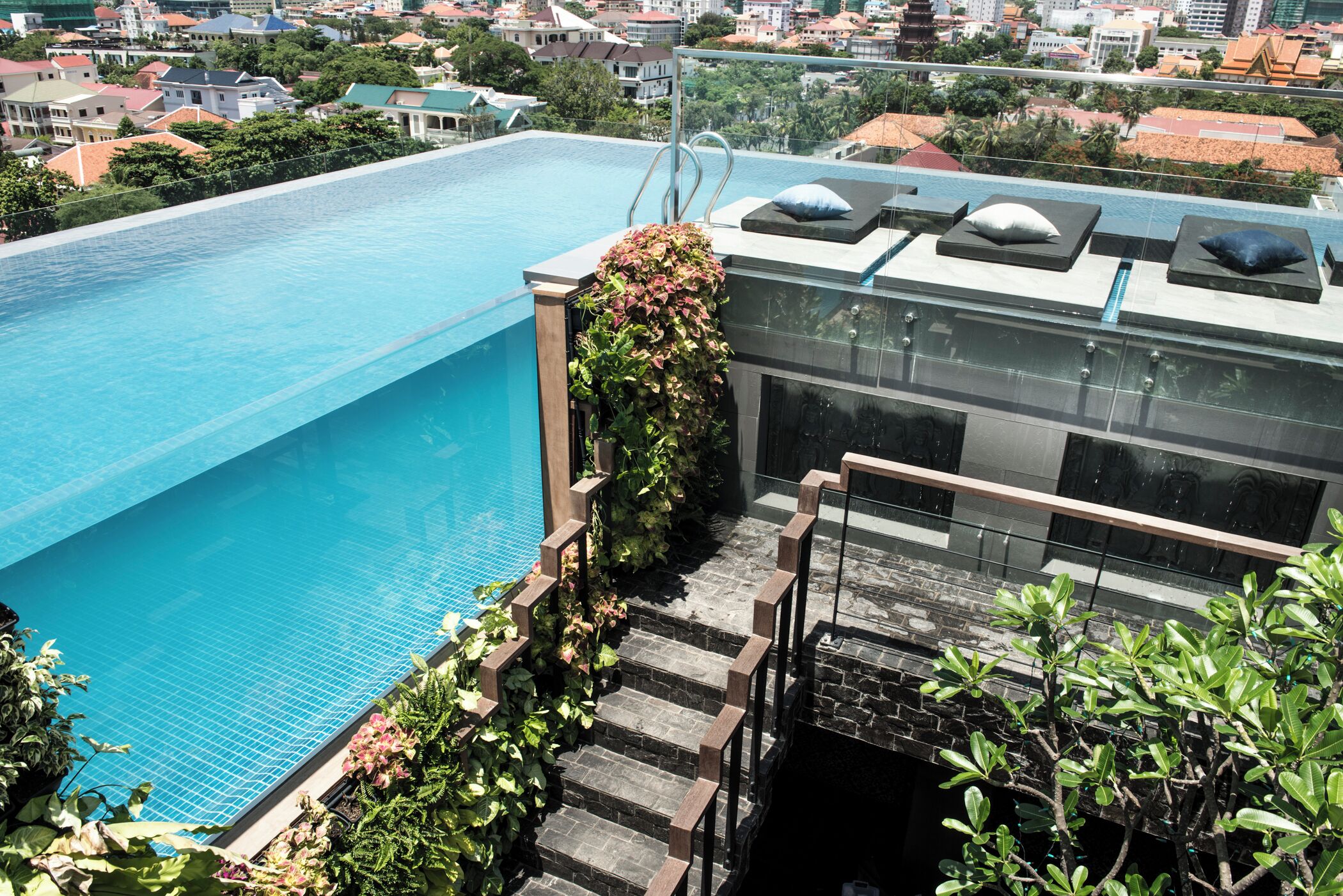 A rooftop infinity pool with a glass balustrade, surrounded by lush greenery and a cityscape view.