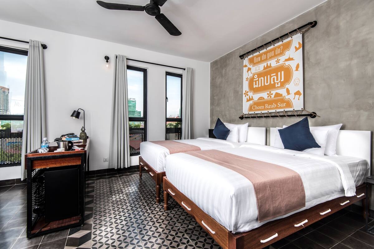 A hotel room with two beds, a desk with a lamp, a ceiling fan, and a large window with curtains.