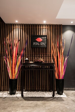 Ett väntrum med en skylt som visar texten ”PETAL SPA”, en vägg med träribbor, en receptionsdisk och två stora krukväxter med färgstarka blad.