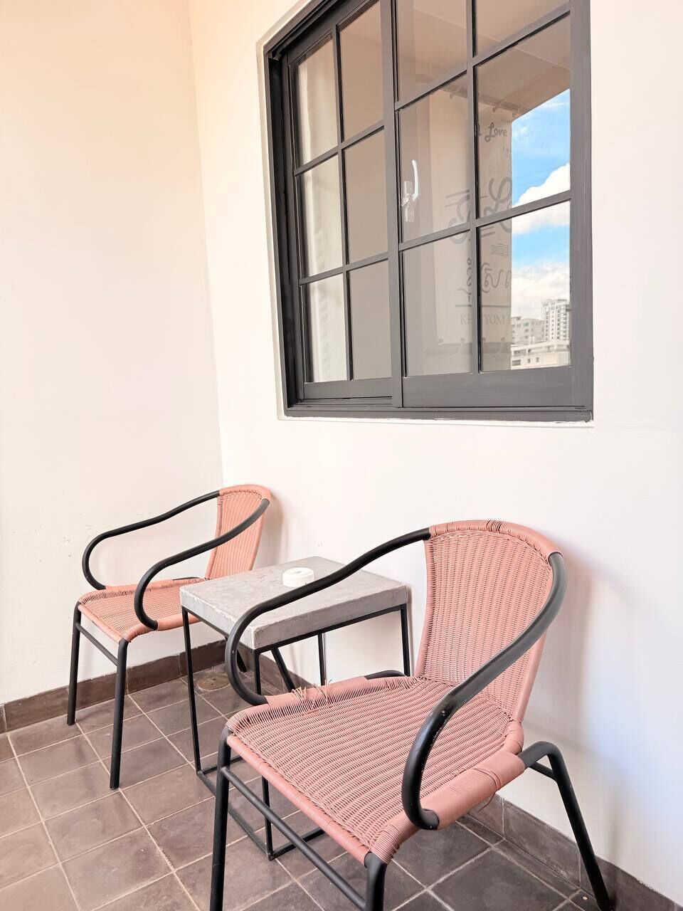 A corner room with two wicker chairs and a small table, a window with a view of buildings, and a tiled floor.