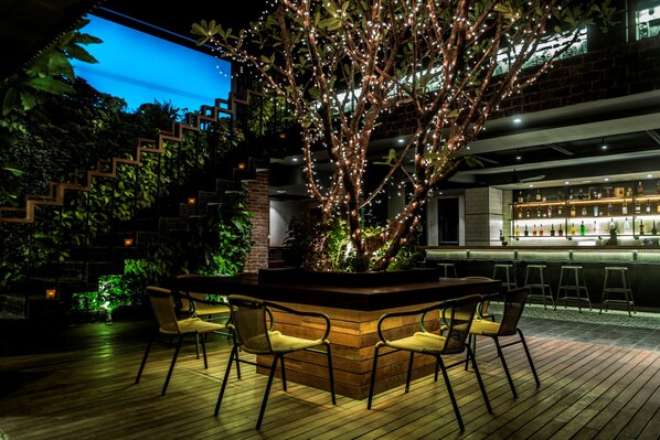 Zona de bar al aire libre con una mesa y sillas de madera, rodeada de follaje e iluminada con luces colgantes.
