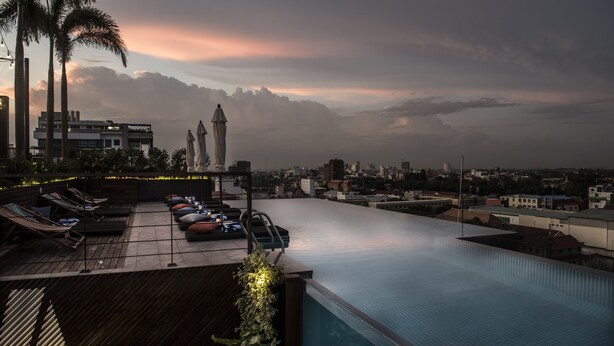 An infinity pool with lounge chairs and umbrellas, overlooking a cityscape with mountains in the background. - Aquarius Hotel & Urban Resort Phnom Penh (Phnom Penh)
