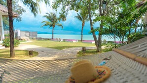 Una hamaca con un sombrero de paja sobre un tapete tejido, con vista a una zona de piscina, palmeras y el mar.