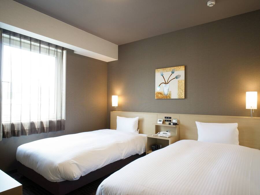 A hotel room with two beds, a bedside table with a lamp, a clock, and a painting on the wall.