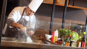 A chef in a white uniform and hat preparing food at a counter with various condiments and fresh herbs.
