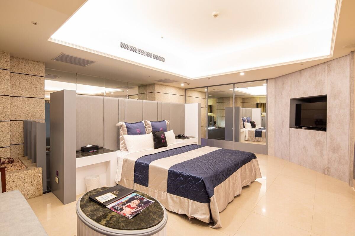 A modern hotel room with a bed, a small round table with a magazine, a TV, and a large mirror.