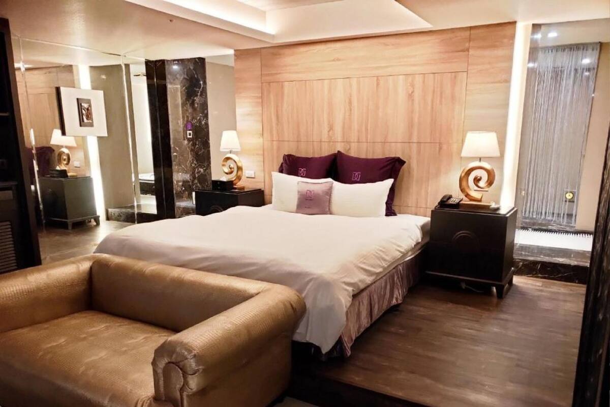 A modern hotel room with a large bed, a leather armchair, and a wooden headboard.