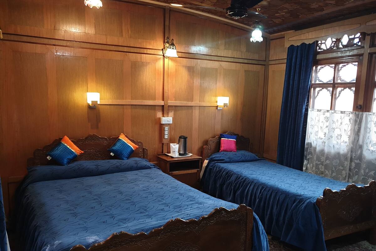 Quarto com duas camas de madeira, cada uma com colcha azul e almofadas decorativas. Há luminárias fixadas na parede e uma janela com cortinas.