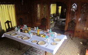Mesa de jantar posta com pratos, talheres e diversas bebidas, rodeada por mobiliário de madeira e carpete estampado.