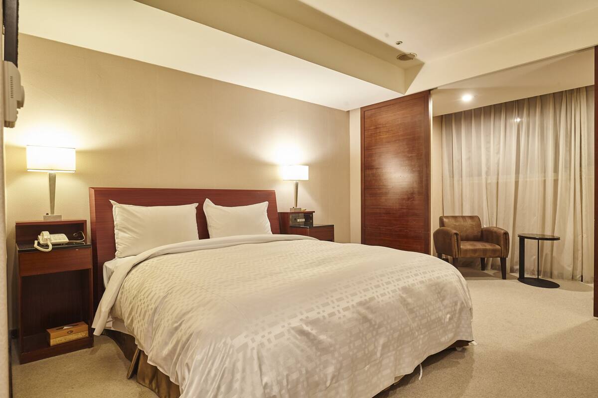 A hotel room with a large bed, two bedside lamps, a wooden headboard, a chair, a small table, and a curtain-covered window.