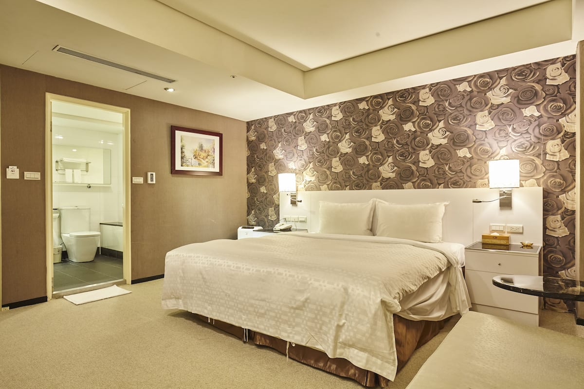 A hotel room with a large bed, a nightstand, a bedside table, a wall with a floral pattern, and a bathroom visible through an open door.