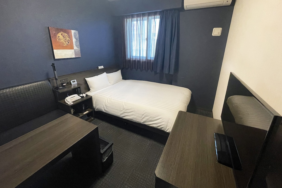 A hotel room with a bed, a desk, a chair, a nightstand, a telephone, and an air conditioning unit.