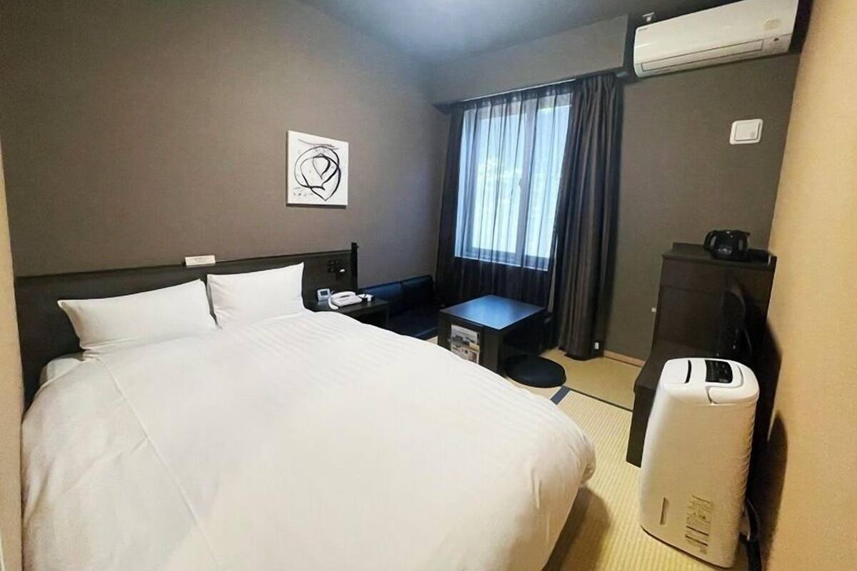 A hotel room with a large bed, a desk, a chair, a small table, an air conditioning unit, and a window with curtains.