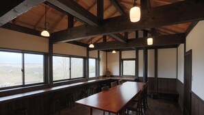 A spacious room with a long wooden table, multiple chairs, and large windows offering a view of the outdoors. - Sun & Moon Club (Fujinomiya)