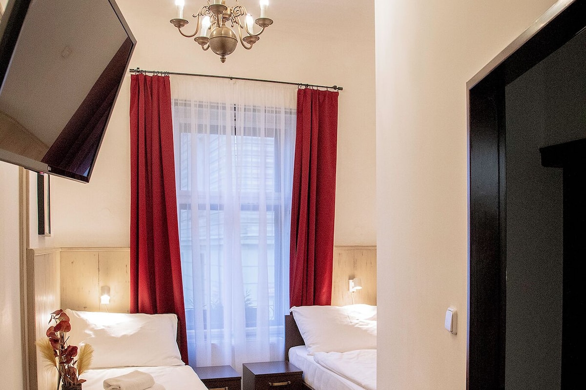 A hotel room with two beds, a TV mounted on the wall, a small table, two red chairs, and a chandelier.