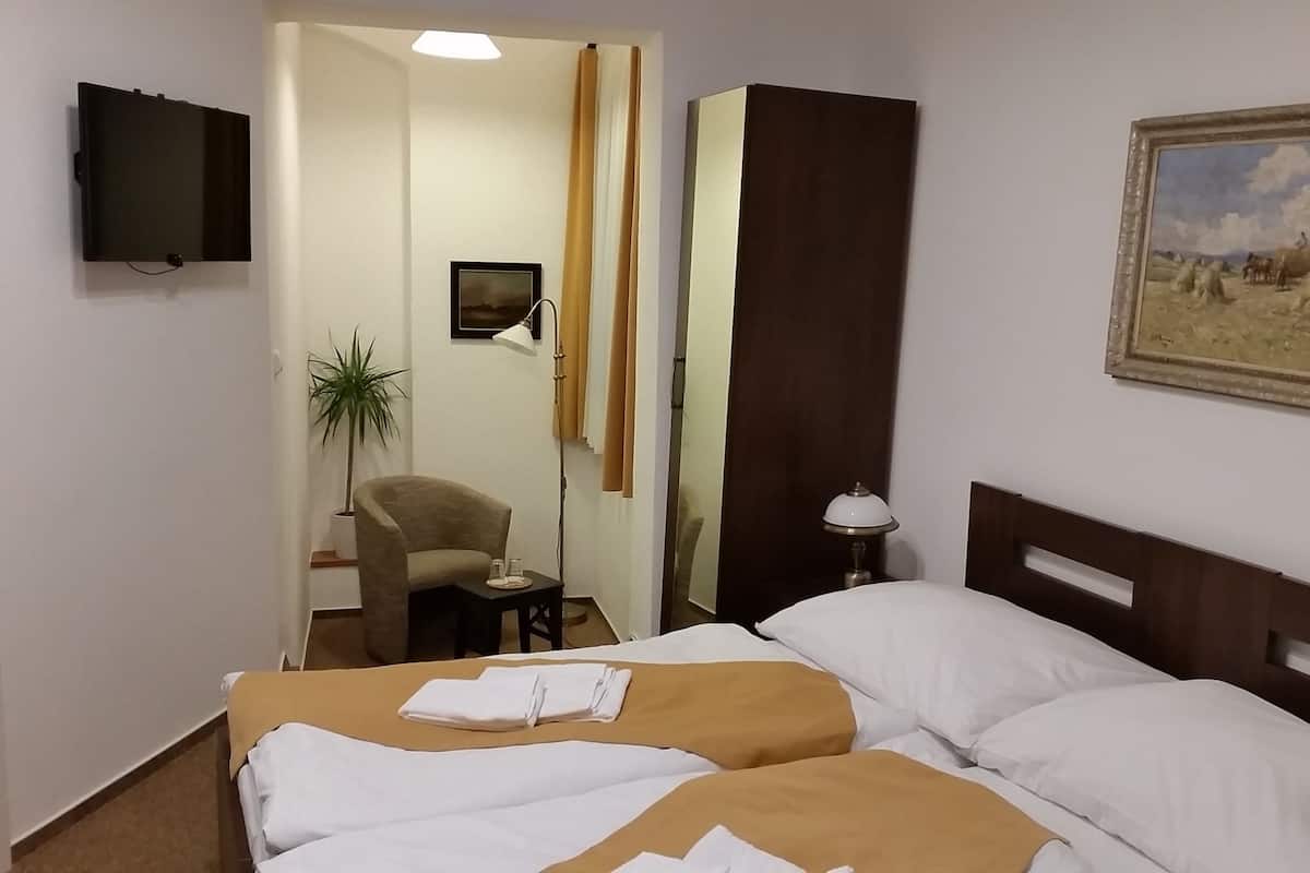A hotel room with two beds, a chair, a desk, a TV, a painting, and a plant.
