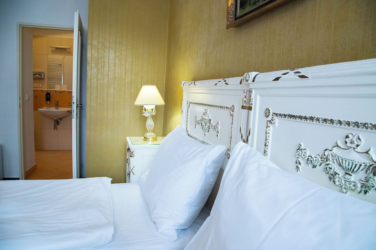 A hotel room with a bed, a nightstand with a lamp, a bathroom visible through an open door, and a wall-mounted picture frame.