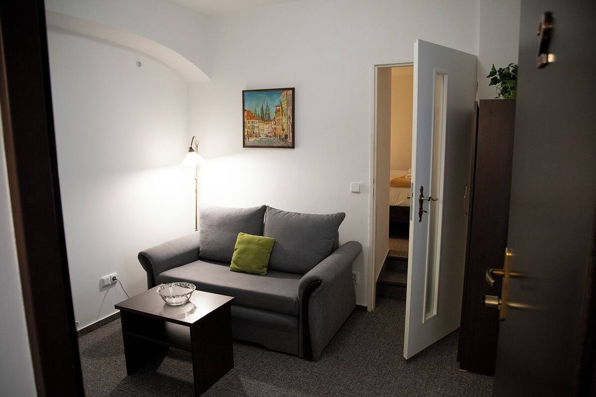 A small, furnished room with a grey sofa, a small table with a glass bowl, a painting on the wall, and a door leading to another room.