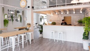A modern cafe with a high ceiling, white tiled walls, and a wooden counter. There are white stools and a long wooden table. - Movylodge Hostel (Bangkok)