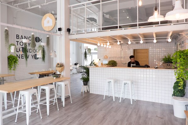 Un café moderno con techo alto, paredes de baldosas blancas y una barra de madera. Hay taburetes blancos y una mesa larga de madera.