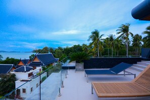A rooftop terrace with a view of the sea, palm trees, and traditional buildings.