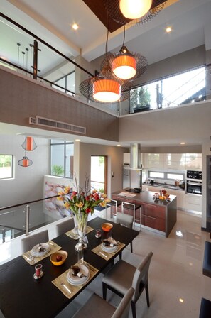 A modern kitchen with a central island, a dining area with a long table, and a staircase leading to an upper level.