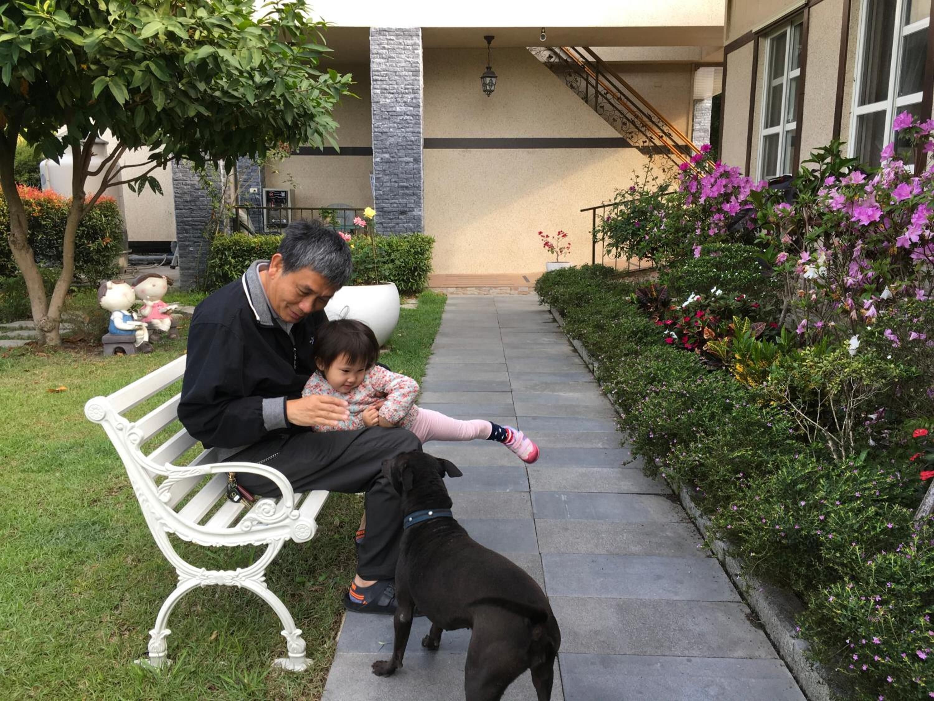 A man sitting on a bench with a child and a dog in a garden area with a pathway and flower beds.