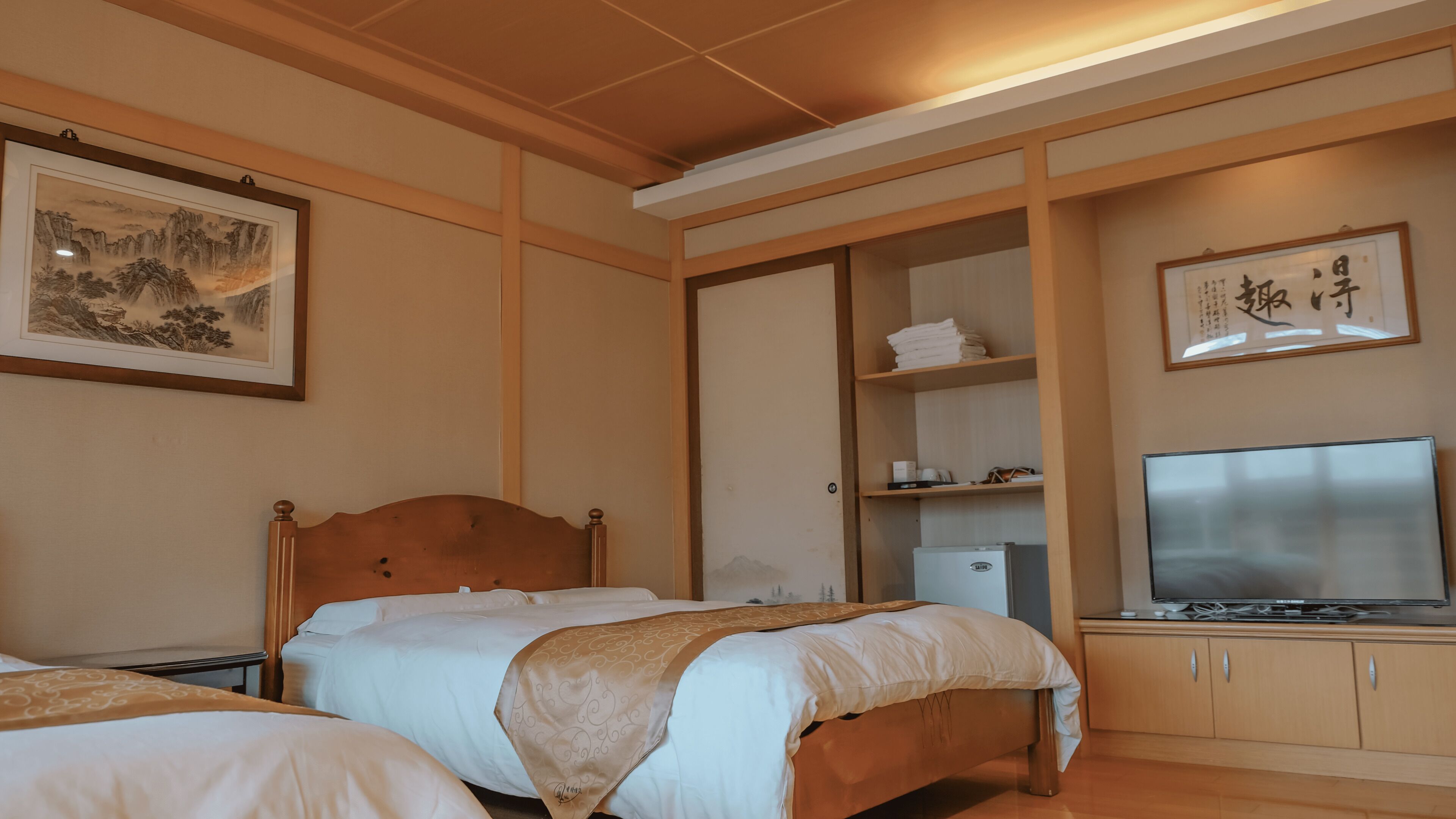 A hotel room with a bed, a TV, a wardrobe, and a shelf with towels and a small refrigerator.