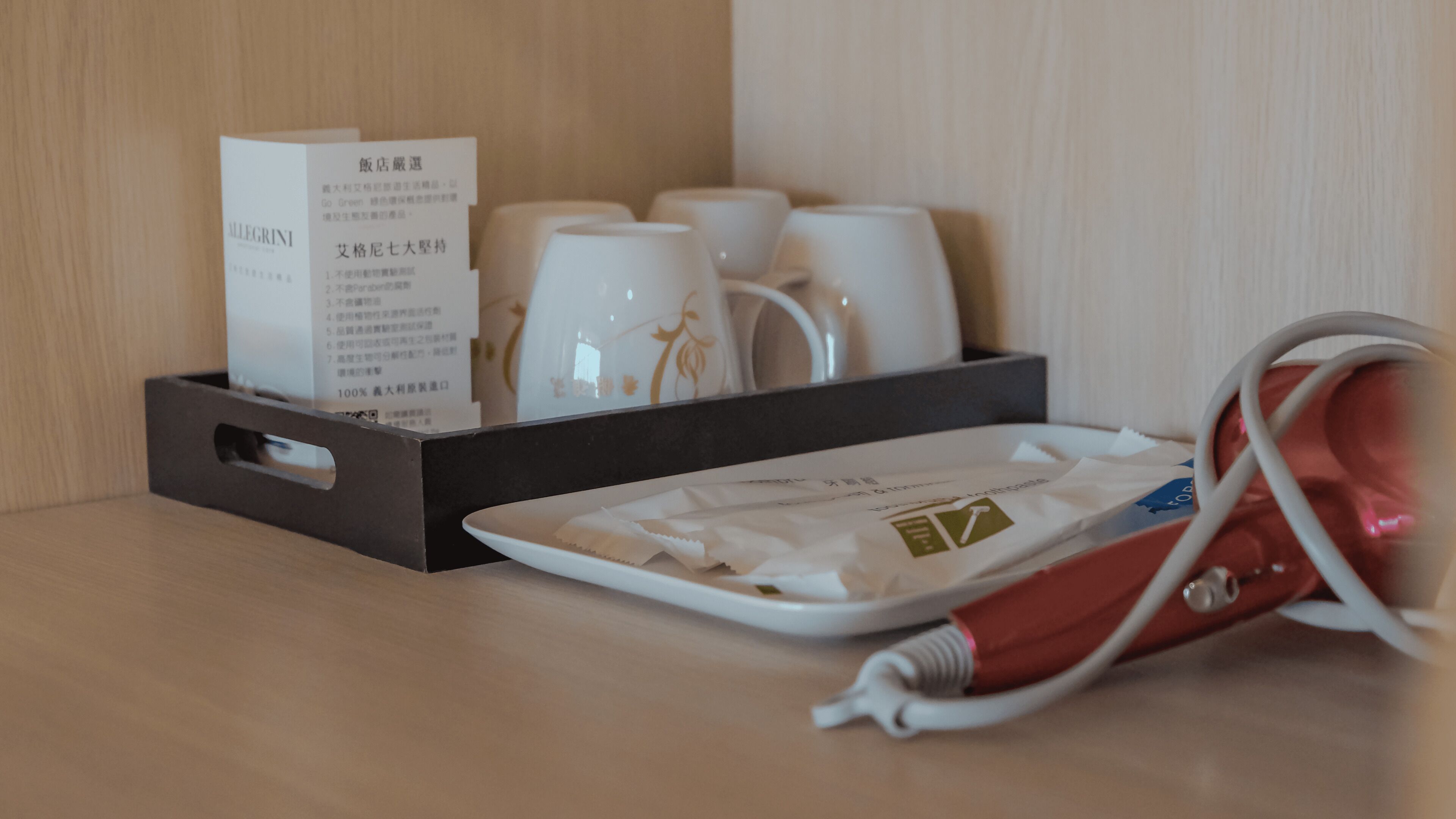 Hotelkamer met een serveerblad waarop een witte mok met gouden decoraties, een zwart blad met een witte beker en een menukaart, en een rood-witte elektrische tandenborstel staan.