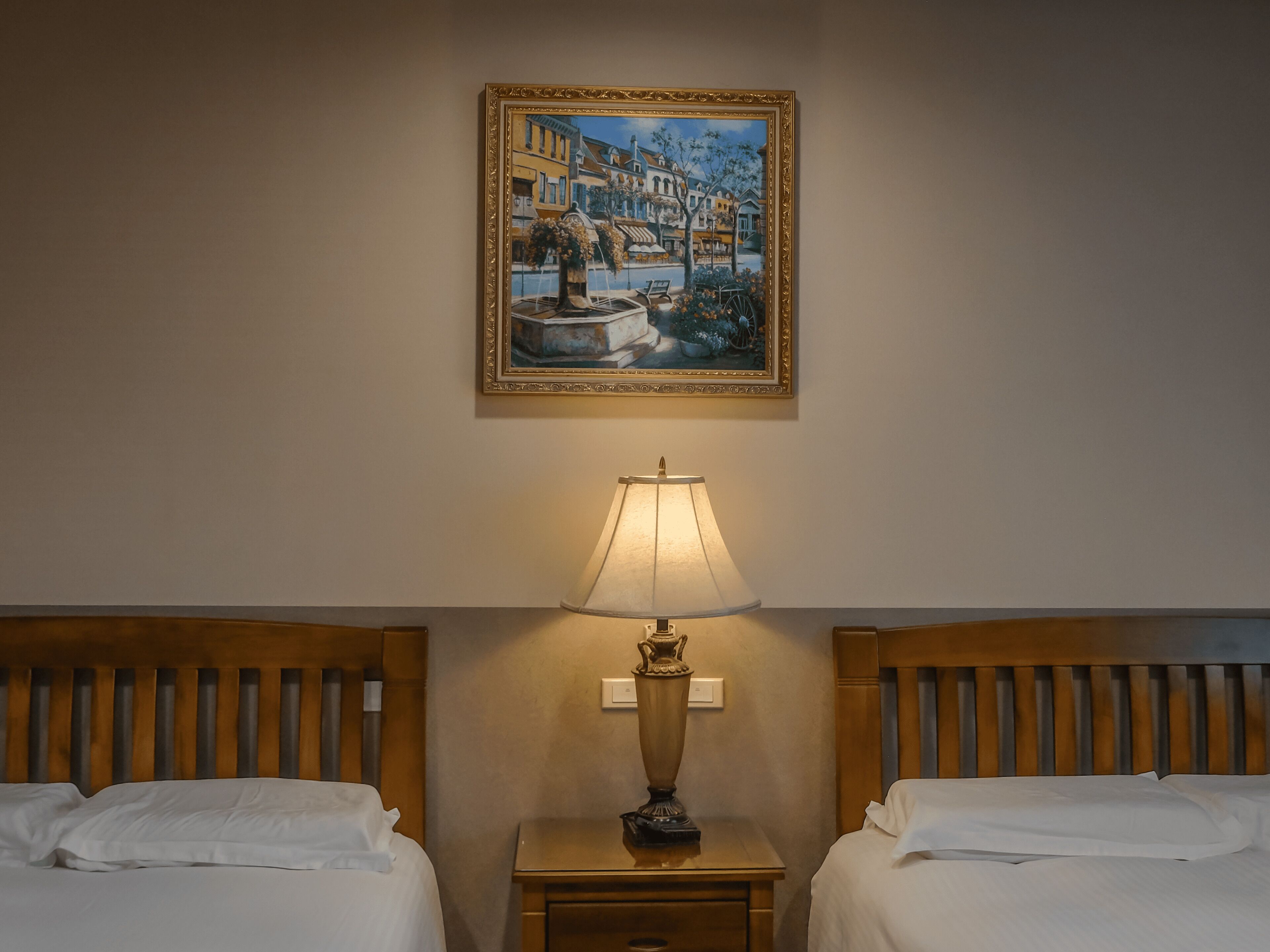 A hotel room with two beds, a nightstand with a lamp, and a painting of a town scene on the wall.