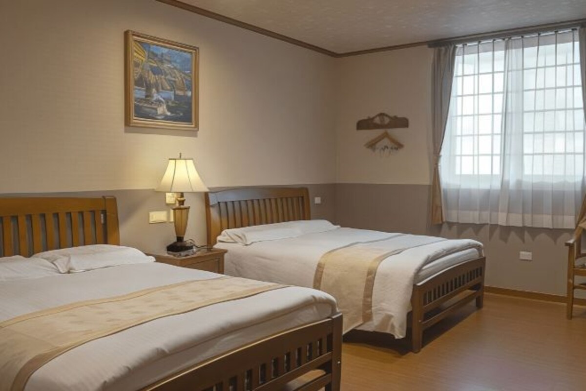 A hotel room with two wooden beds, a nightstand with a lamp, a painting on the wall, and a window with curtains.