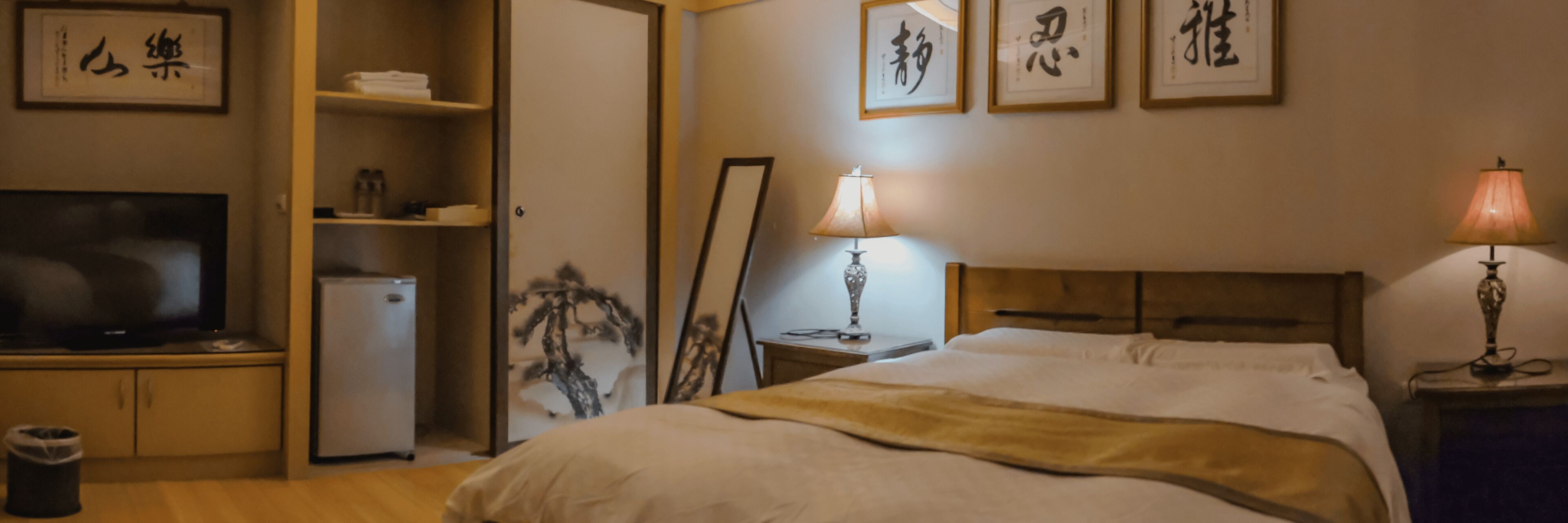 A hotel room with a bed, a television, a refrigerator, and framed calligraphy on the wall.