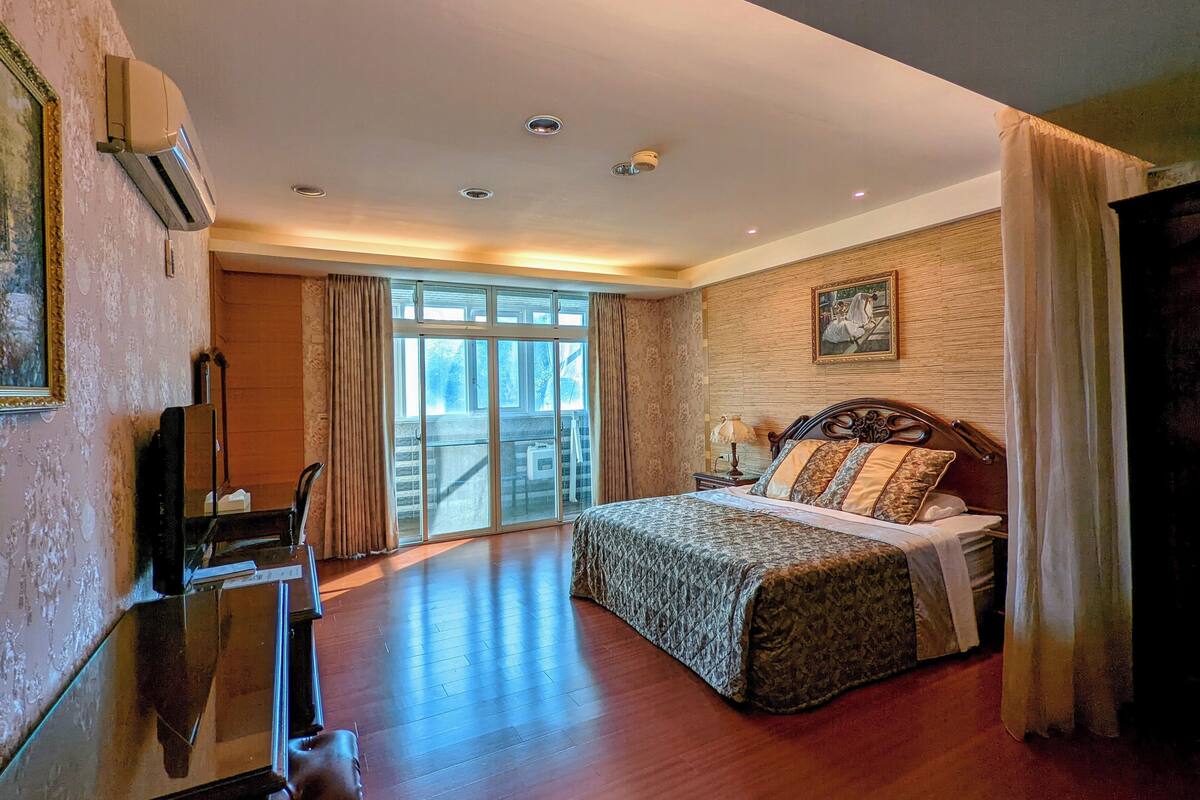 A spacious bedroom with a large bed, a sliding glass door to the balcony, and a ceiling-mounted air conditioning unit.
