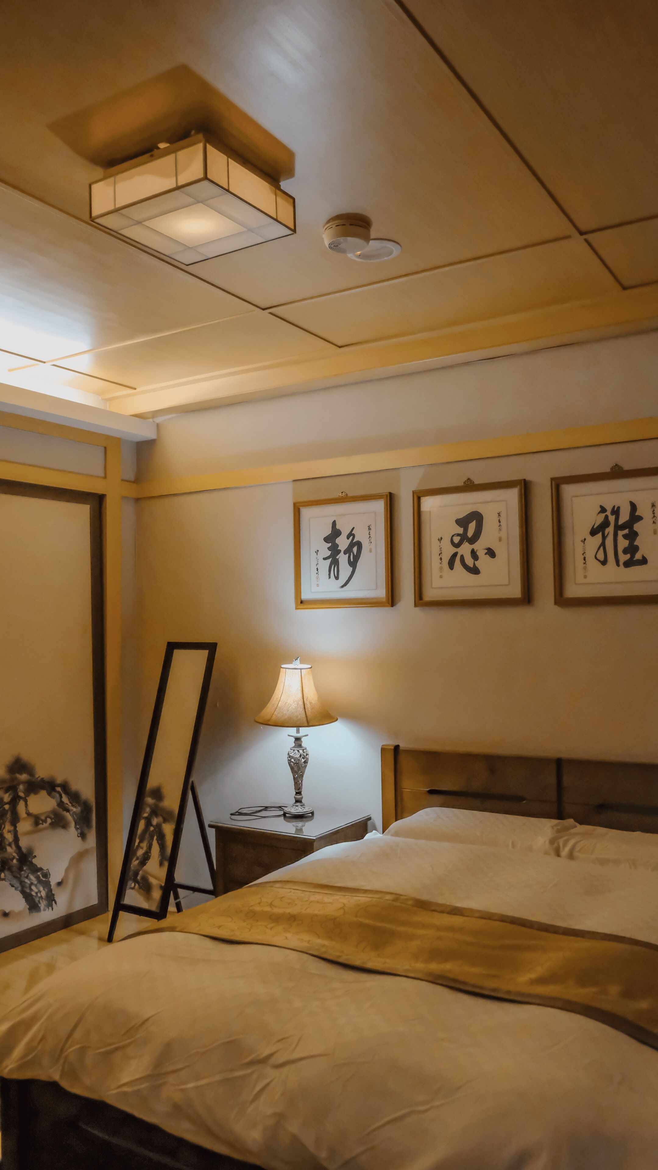 A hotel room with a bed, a nightstand with a lamp, framed calligraphy on the wall, and a ceiling light fixture.