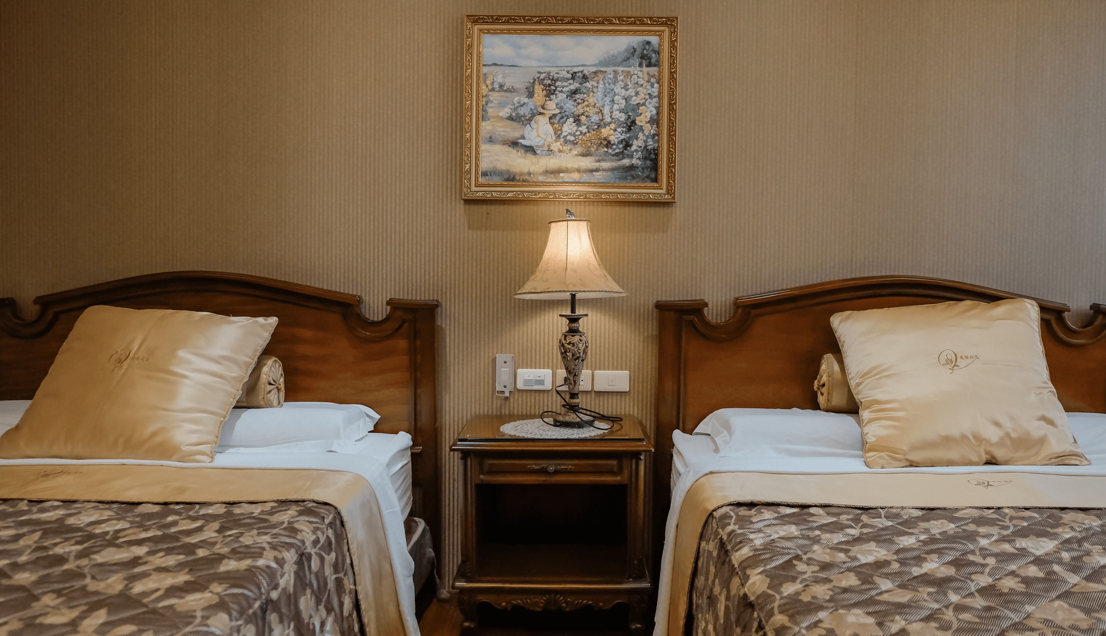 A hotel room with two beds, a nightstand with a lamp, a painting on the wall, and a wooden headboard.