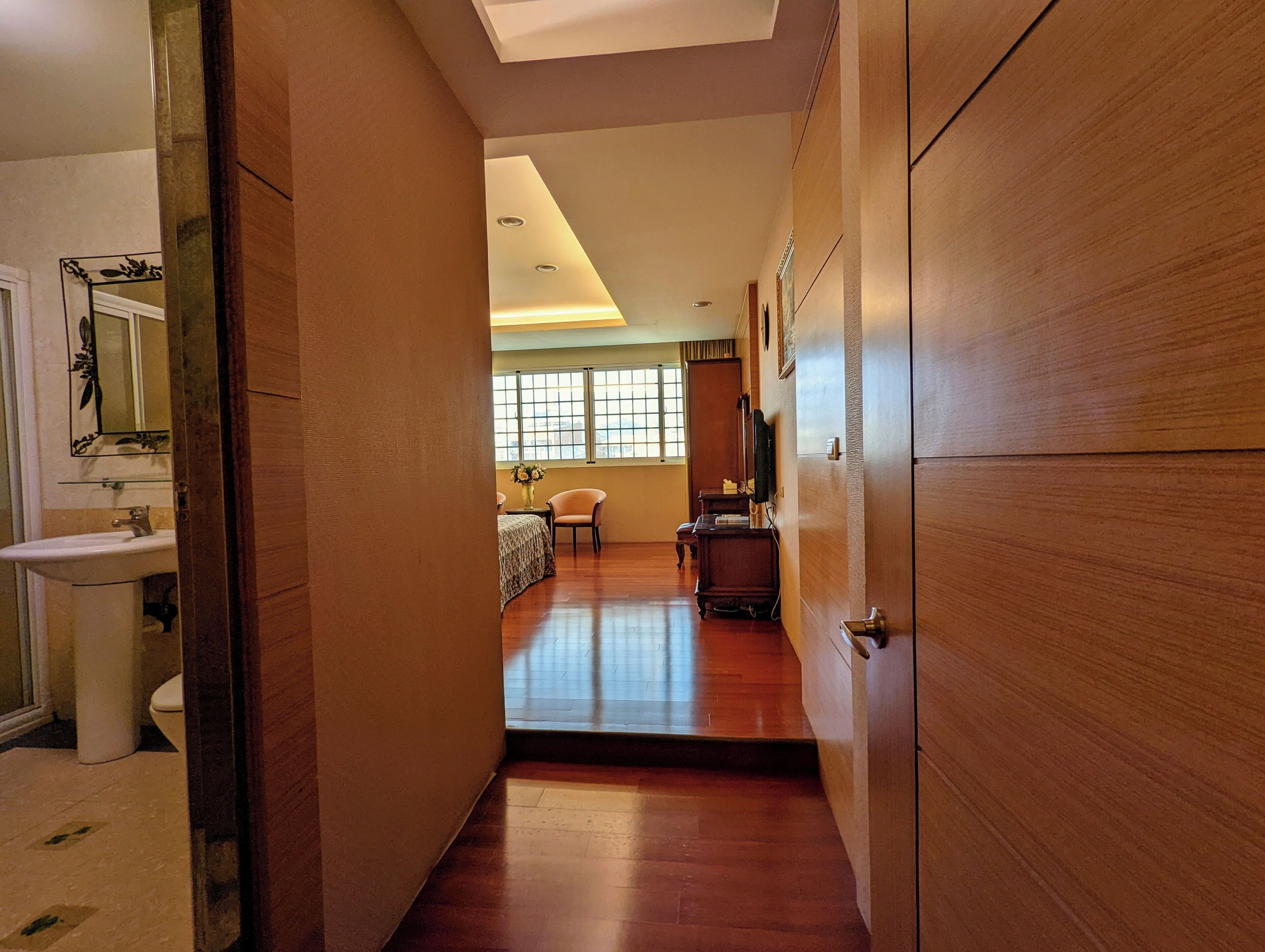 A hallway leading to a bedroom with a wooden floor, a bed, a nightstand, and a window with a view.