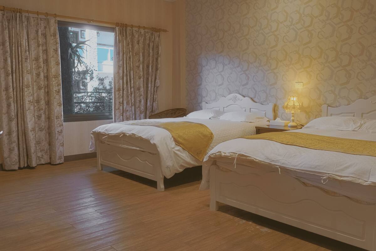 A hotel room with two beds, a window with curtains, a patterned wall, and a bedside lamp.