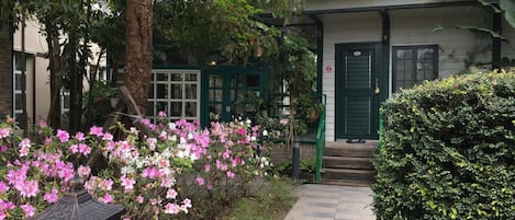 Un edificio con puerta verde y ventanas, rodeado de un jardín con flores rosadas y un sendero de piedra.