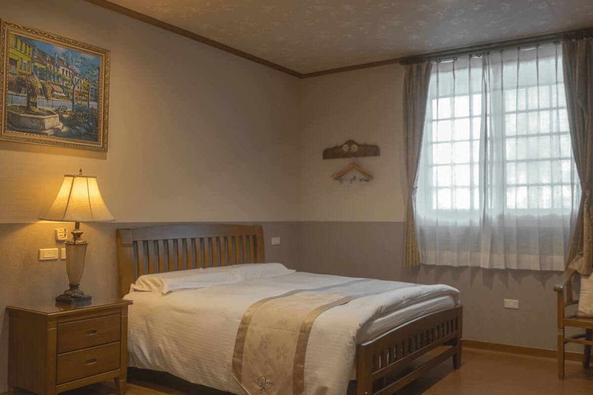 A bedroom with a bed, a nightstand, a lamp, a chair, a window with curtains, and a painting on the wall.