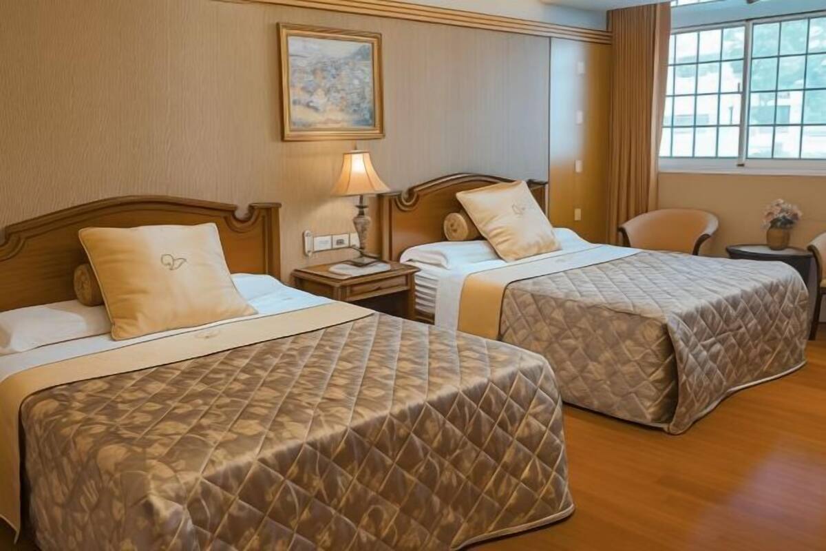 A hotel room with two beds, each with a quilted bedspread and matching pillows. There is a nightstand with a lamp, a painting on the wall, and a window with curtains.