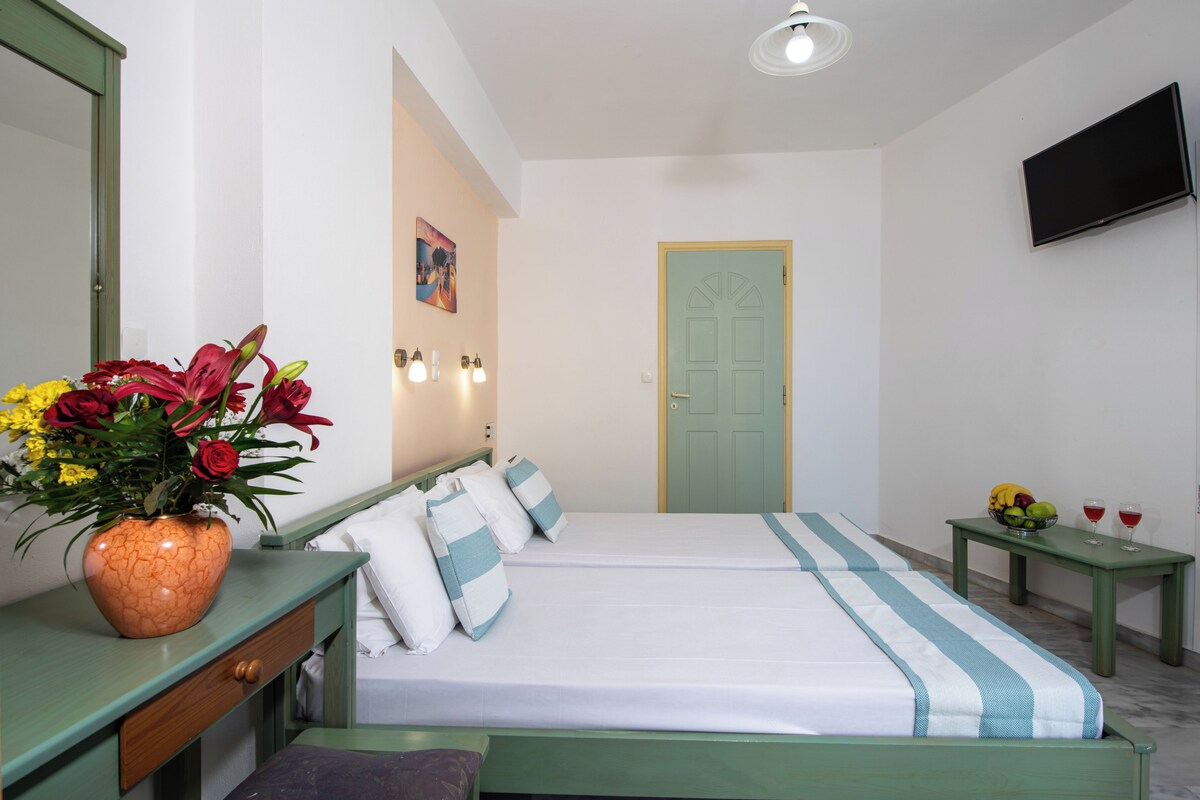 A hotel room with a bed, a bedside table with a vase of flowers, a television mounted on the wall, and a door with a simple design.