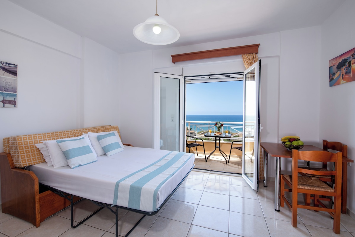 A hotel room with a bed, a small table with a bowl of fruit, a chair, and a view of the sea through an open door.