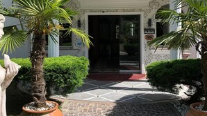 A hotel entrance with a white facade, a balcony with balustrades, and potted plants.