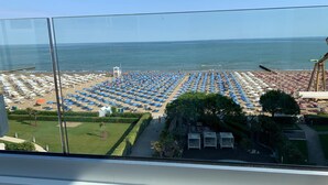 A beach with rows of umbrellas and sun loungers, a clear view of the ocean, and a grassy area with a few trees.